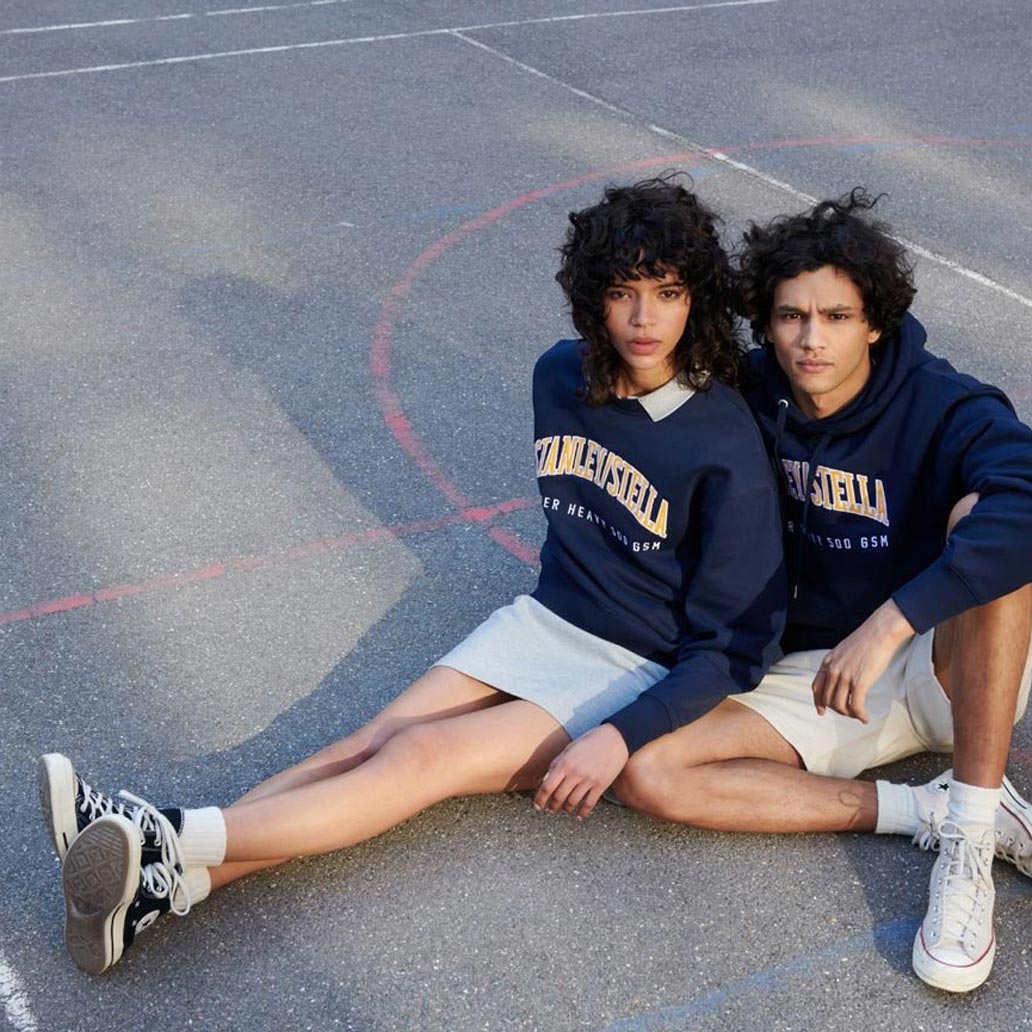 Zwei Menschen sitzen auf einem asphaltierten Basketballplatz und tragen jeweils ein Sweatshirt und einen Hoodie mit Stanley/Stella Aufschrift