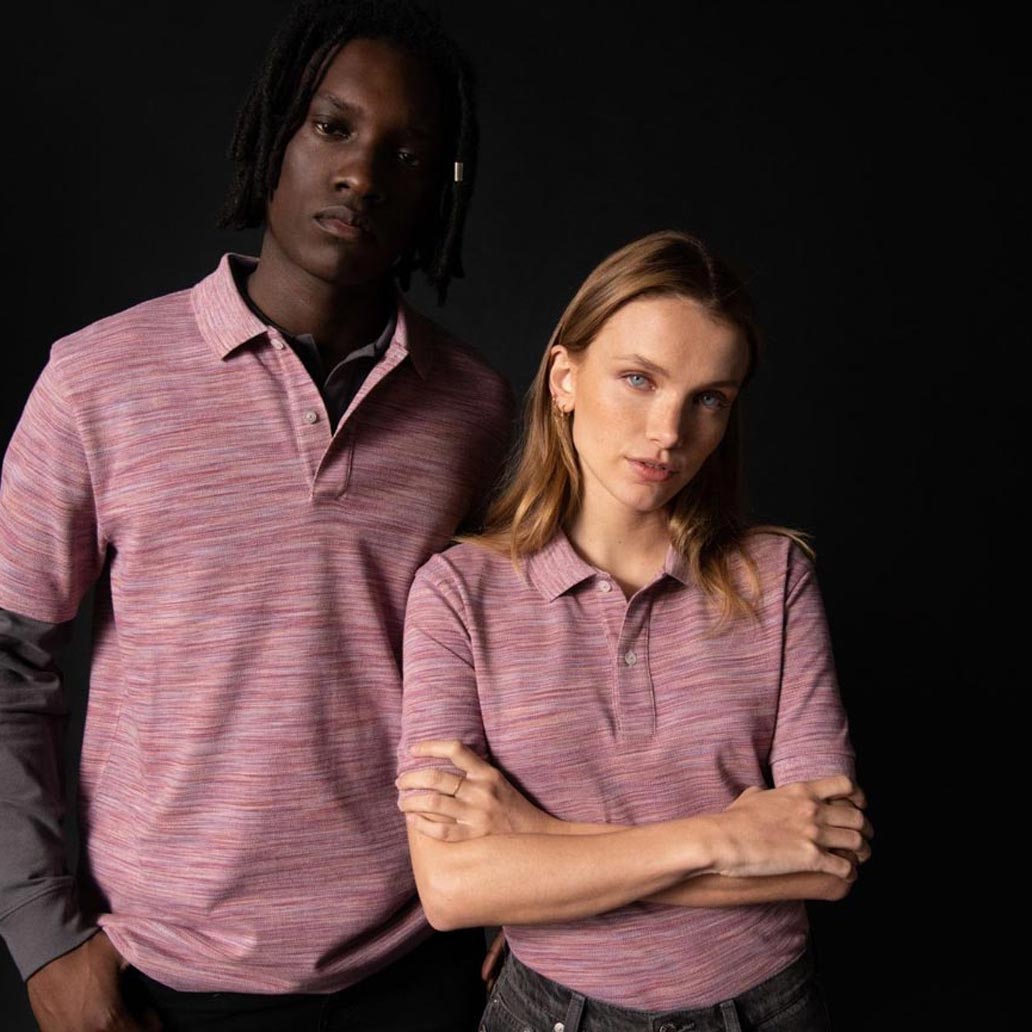 Zwei Menschen stehen vor einem dunklen Hintergrund und tragen rose gestreifte Poloshirts