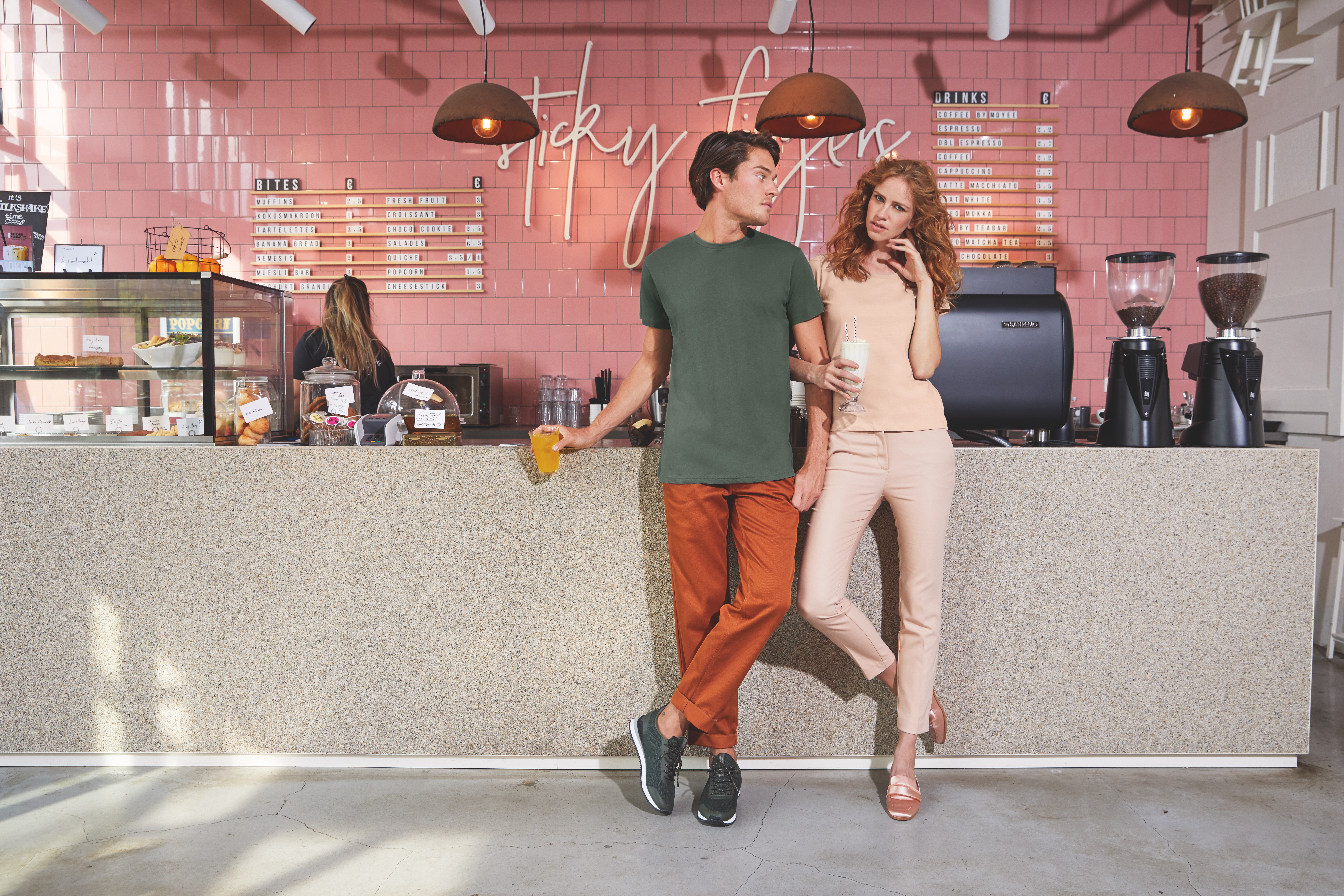 Ein Mann und eine Frau stehen in einem Cafe an der Bar. Im Hintergrund sind rosa Kacheln mit dem Titel "sticky fingers". Beide halten ein Getränk in der Hand.
