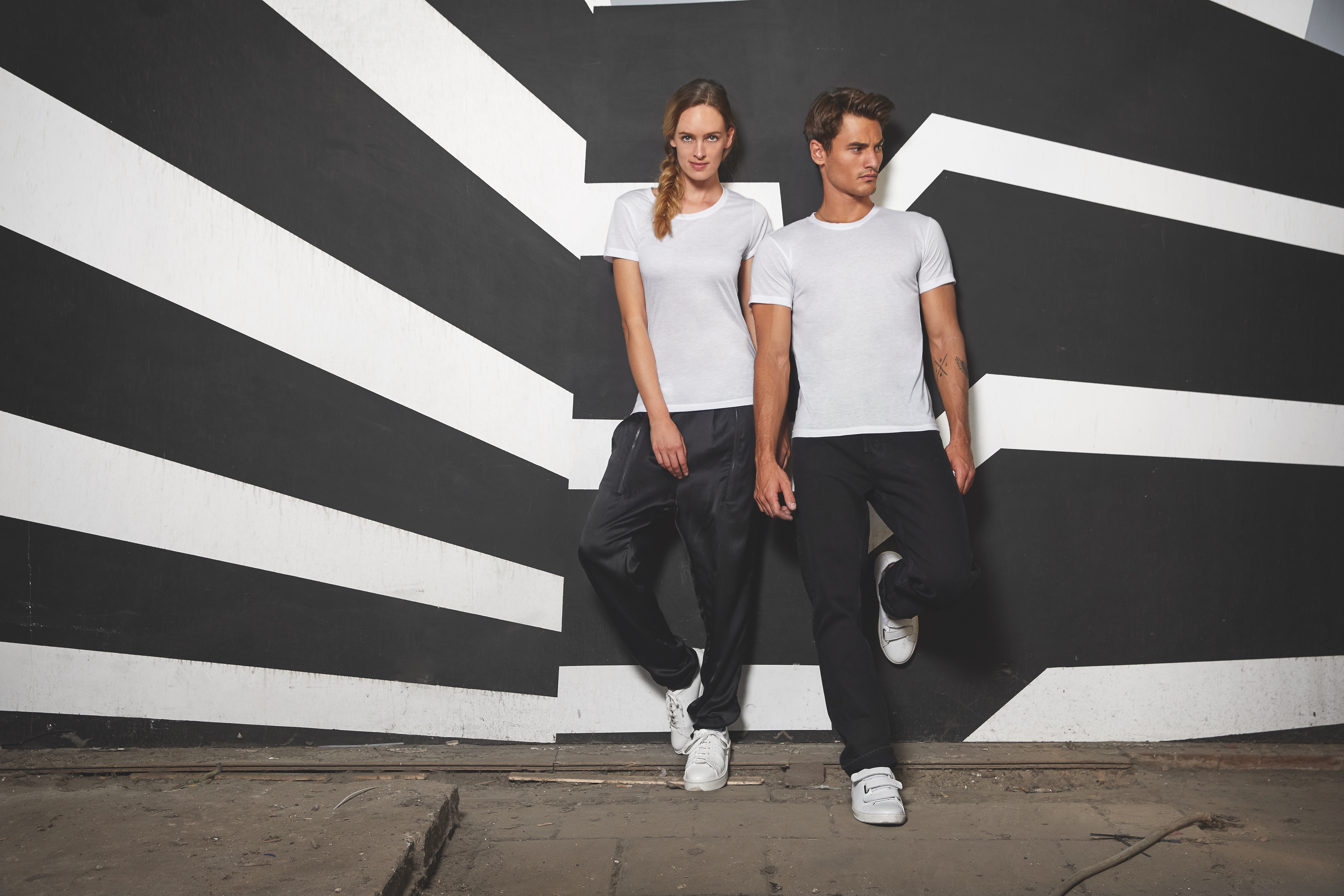 Zwei Menschen in weoßen Shirts und schwarzer Hose vor Schwarz/Weiß gestreiftem Hintergrund